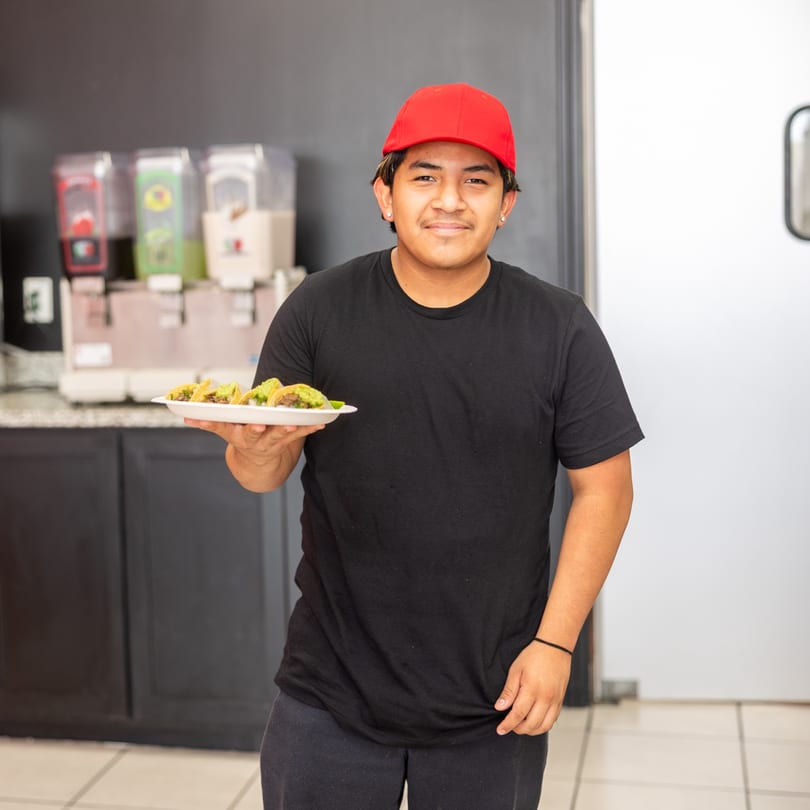 A smiling young man in a red cap and black shirt holds a plate of tacos.