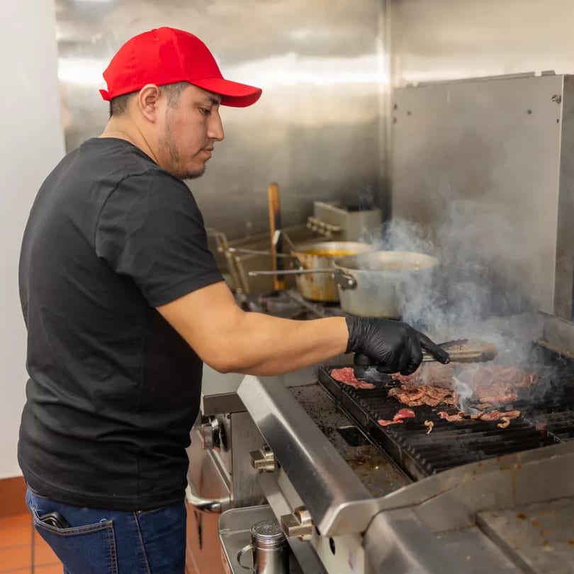 Chef in a red cap and black gloves grilling meat on a smoky griddle in a commercial kitchen.