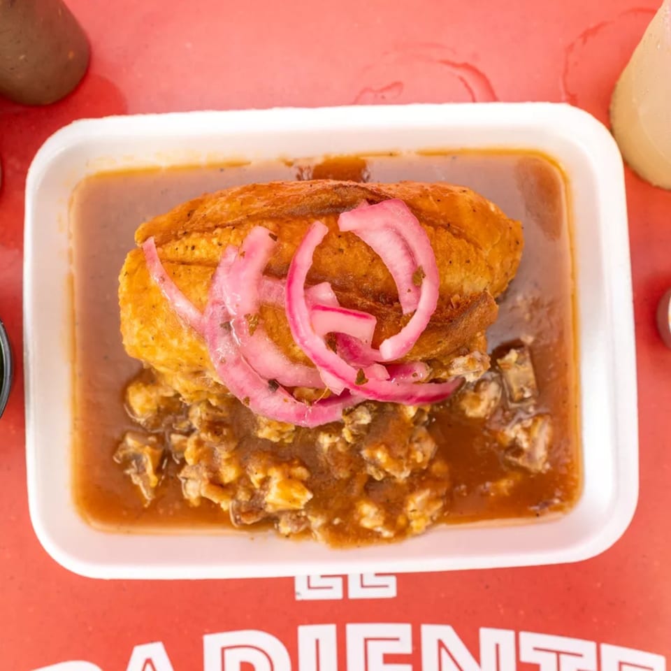A Mexican torta with red pickled onions, submerged in a rich brown sauce, served in a white tray.