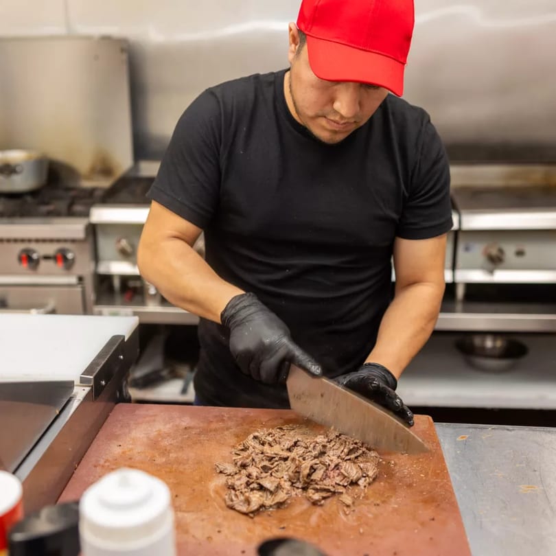 Chef in a red cap and black gloves chopping meat on a cutting board in a commercial kitchen.