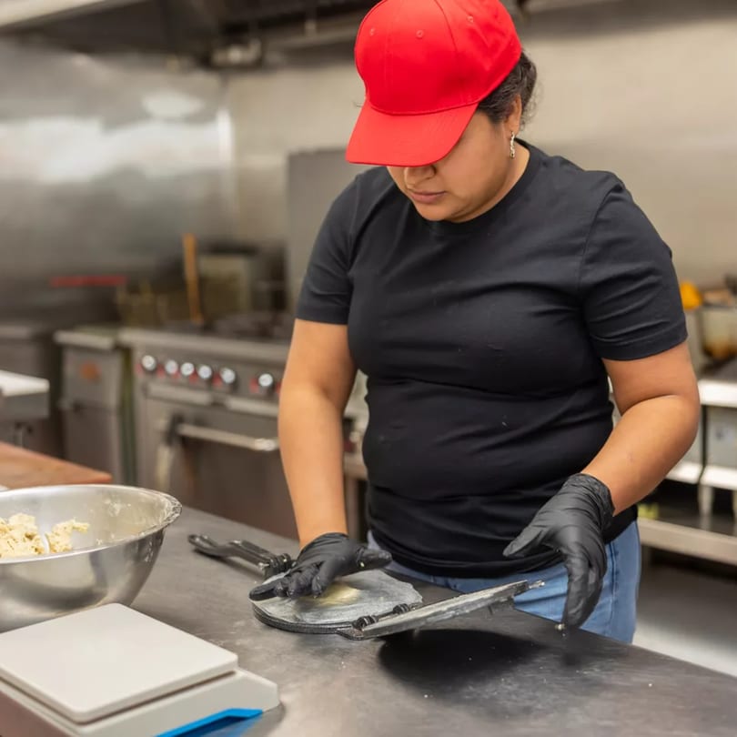 A woman in a red cap and black gloves presses dough in a tortilla maker in a commercial kitchen.