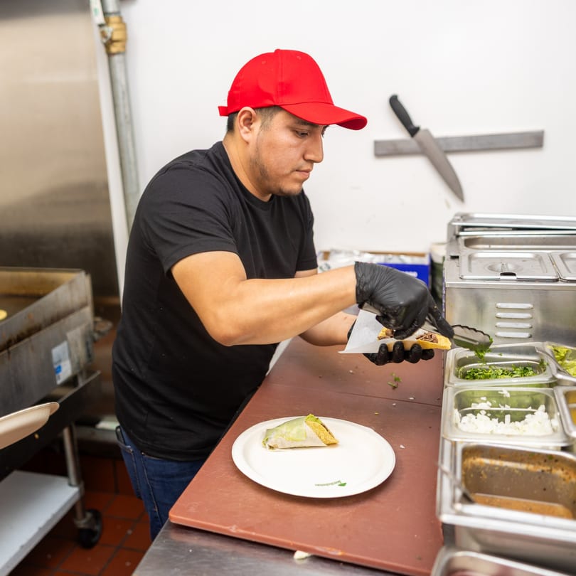 A chef in a red hat and black gloves prepares a taco with fresh ingredients in a commercial kitchen.
