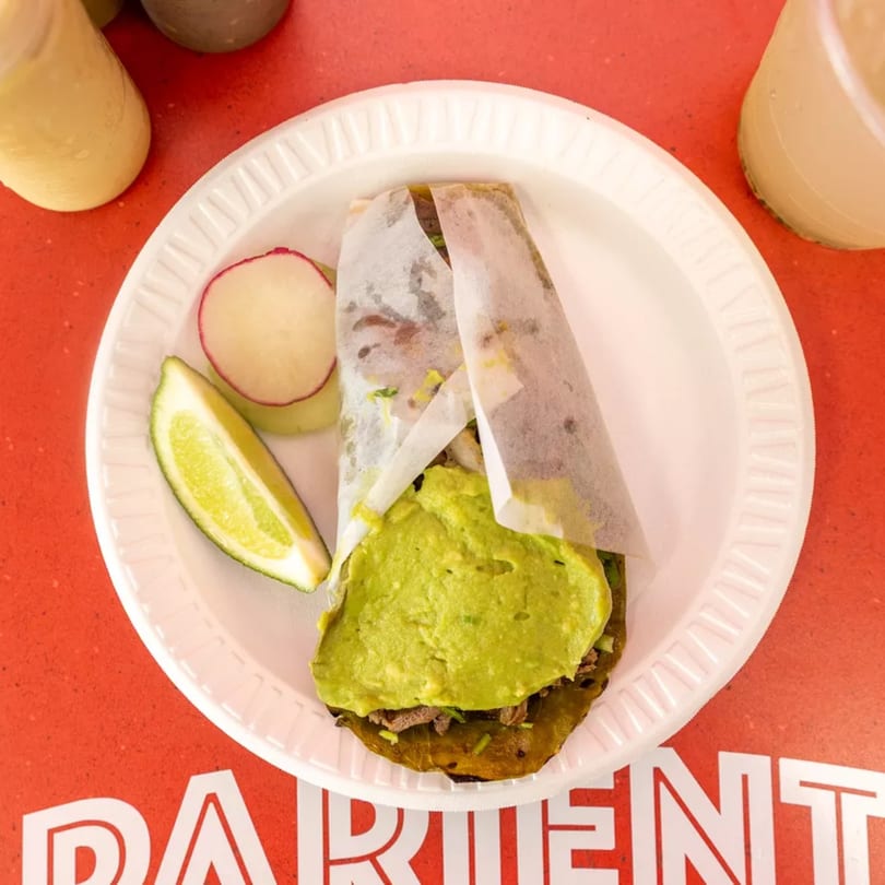 Taco with guacamole, radish, and lime on a white plate, with drinks on a red table.