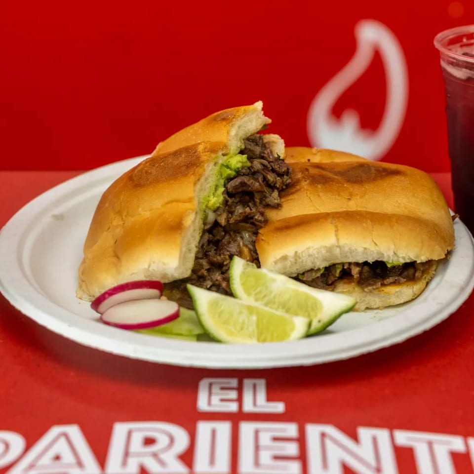 A torta sandwich with meat, avocado, and lime wedges on a white plate with a red background.
