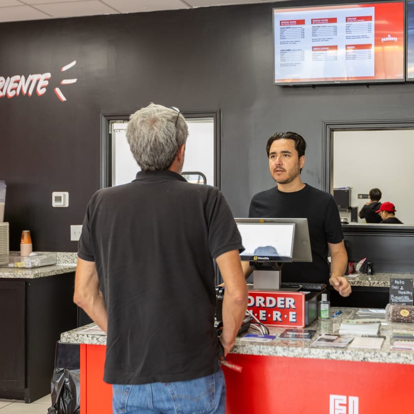 A customer orders at a counter from a male employee, with a menu board visible above.
