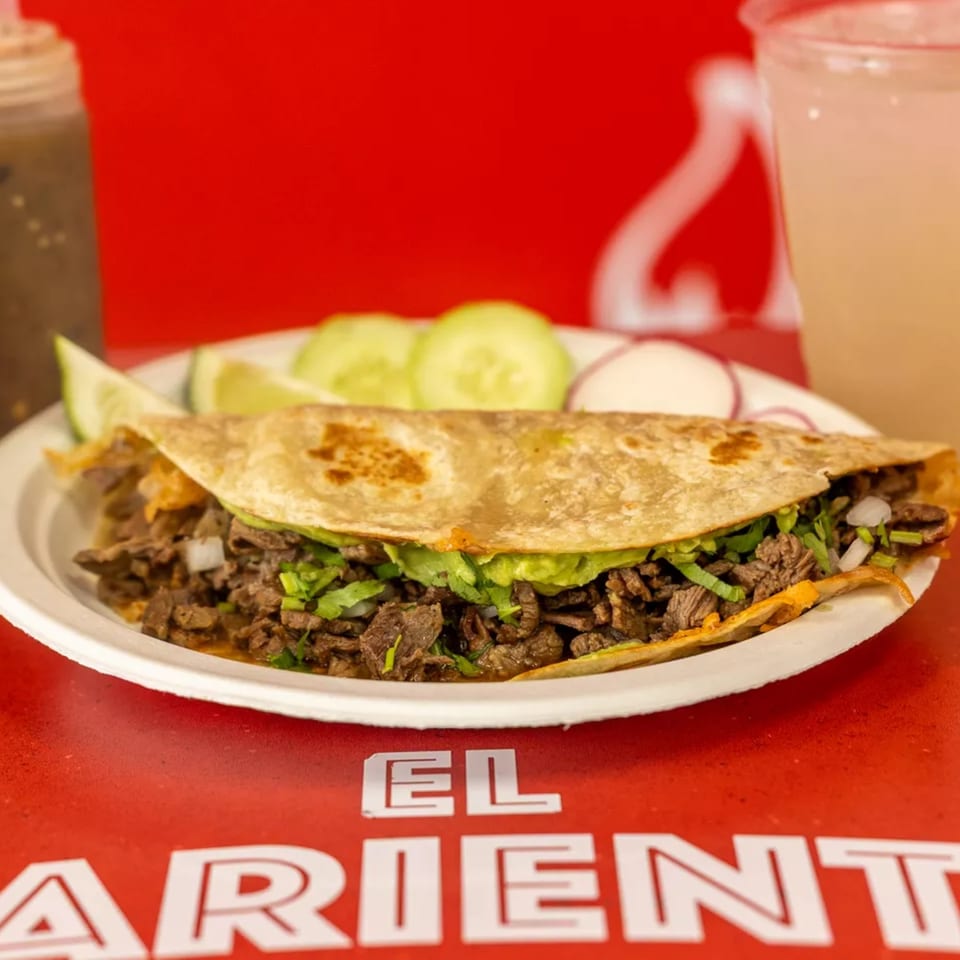 A large quesadilla filled with meat, lettuce, and onions, served with cucumber slices and a drink.